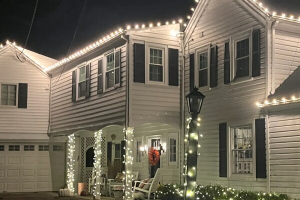 "Beautifully decorated residential home with elegant holiday lighting. Warm white lights outline the roof, wrap around the porch columns, and illuminate the landscaping, creating a cozy and festive atmosphere.