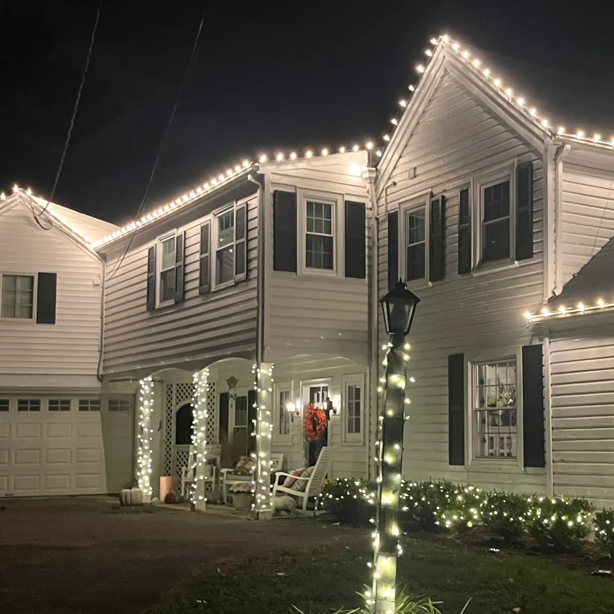 "Beautifully decorated residential home with elegant holiday lighting. Warm white lights outline the roof, wrap around the porch columns, and illuminate the landscaping, creating a cozy and festive atmosphere.